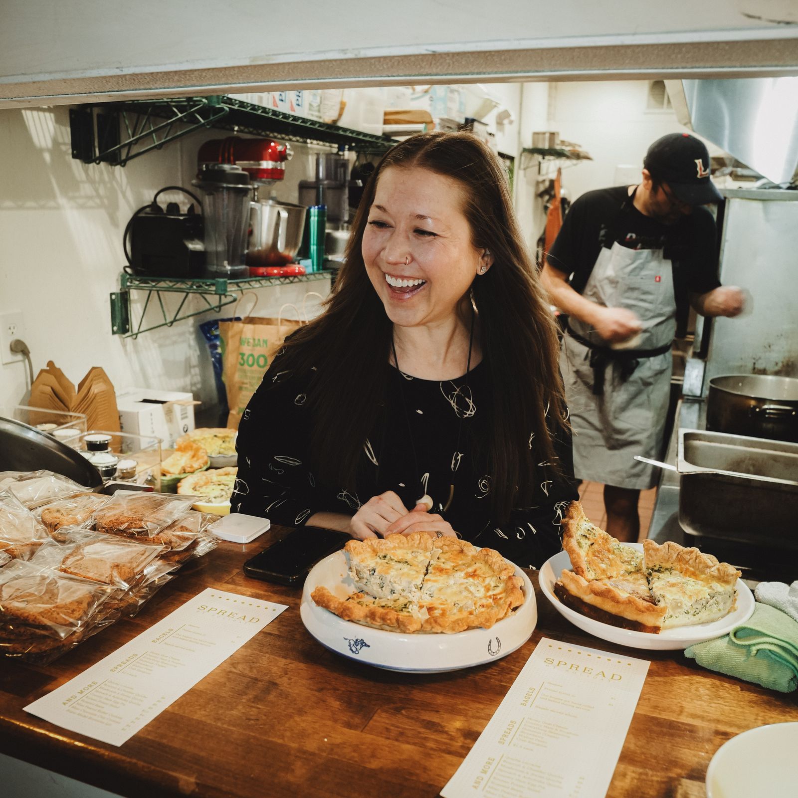 The Spread by Soliday team laughing behind a counter of fresh quiche and baked goods